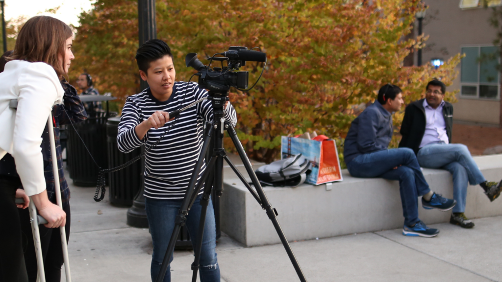 person in striped shirt adjusting a camera positioned on a tripod outside